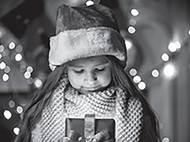 black and white photo of a child holding a christmas gift with christmas lights on in the backround